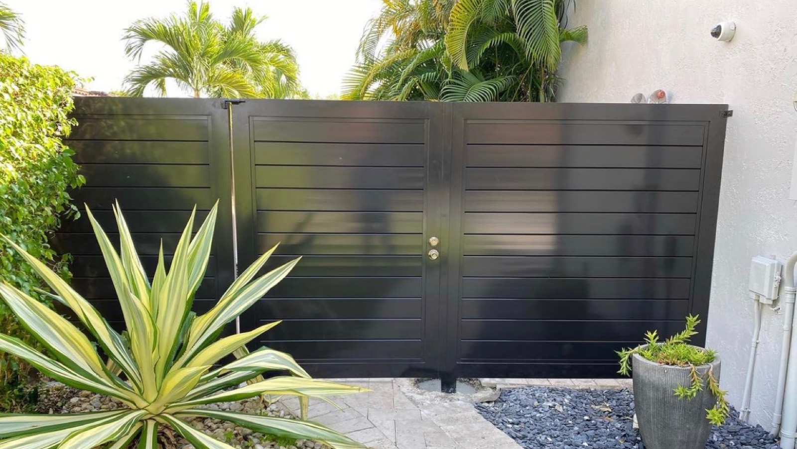 Black horizontal slat driveway gate with tropical landscaping